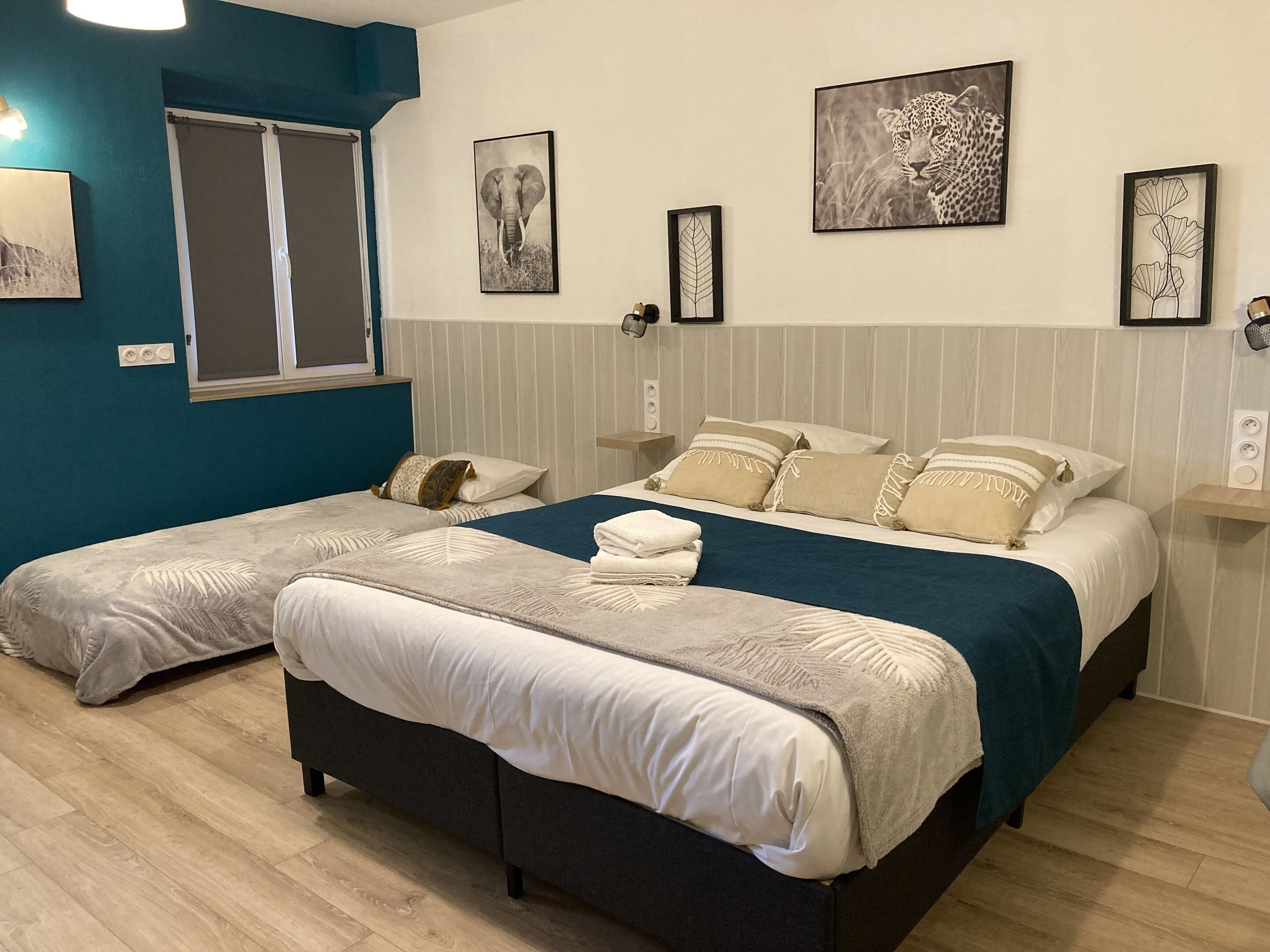 A double bed with a blue headboard, a smaller bed with a white headboard, and a window with blinds.