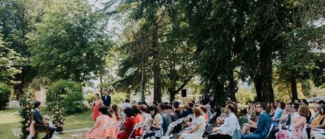 An outdoor event with guests seated on chairs, a stage with a speaker, and trees in the background.