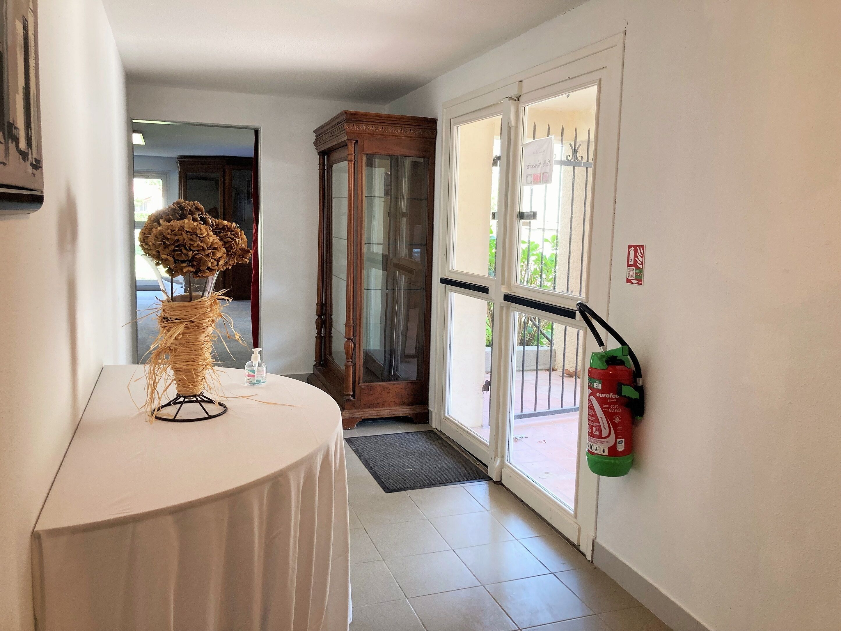 Een hotellobby met een ronde tafel, een vaas met gedroogde bloemen, een brandblusser en een houten kast.