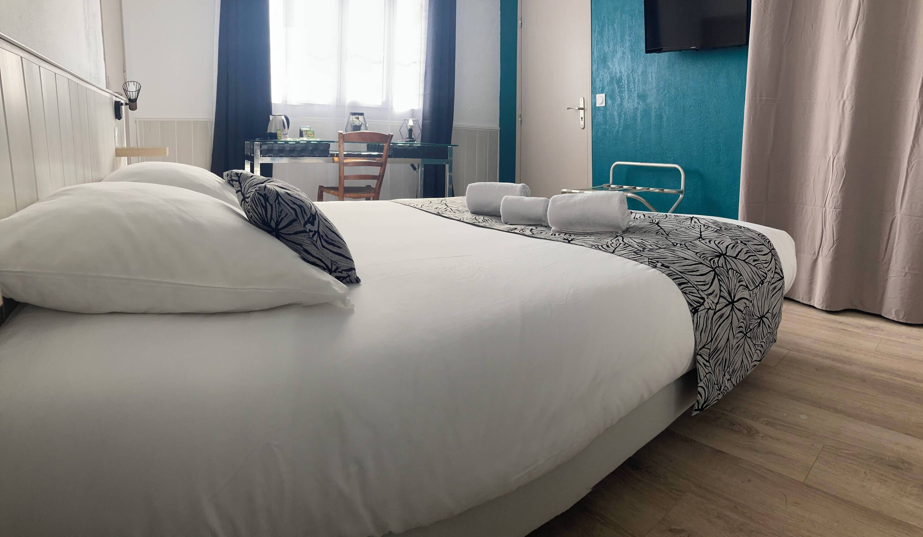 Un lit bien fait, avec des oreillers blancs et à motifs, une table en verre sur laquelle se trouvent une bouilloire et des tasses, une chaise, et un téléviseur fixé au mur, qui est peint dans une nuance de vert turquoise.