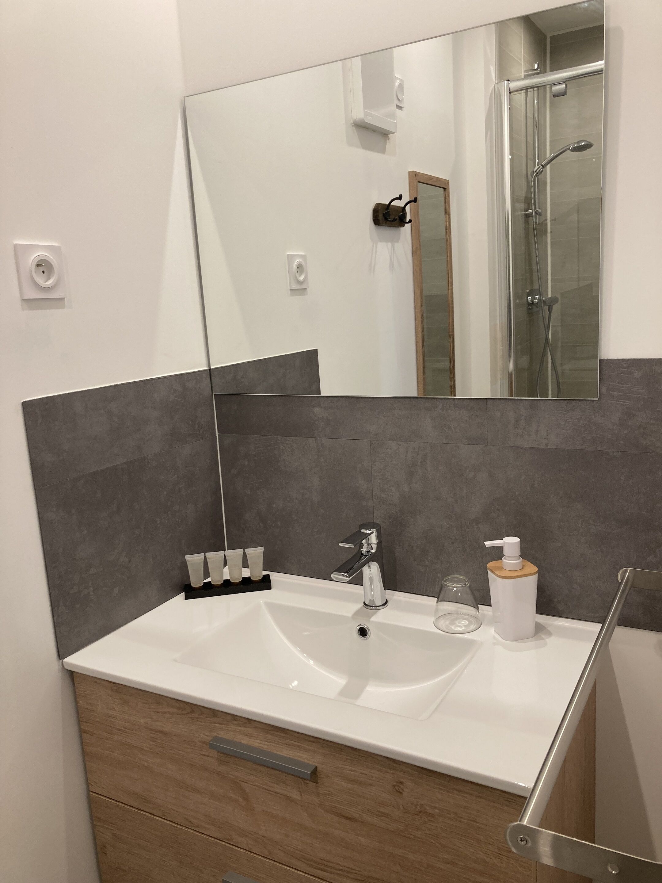 A modern bathroom with a large mirror, a white sink, a shower, and a wooden cabinet.