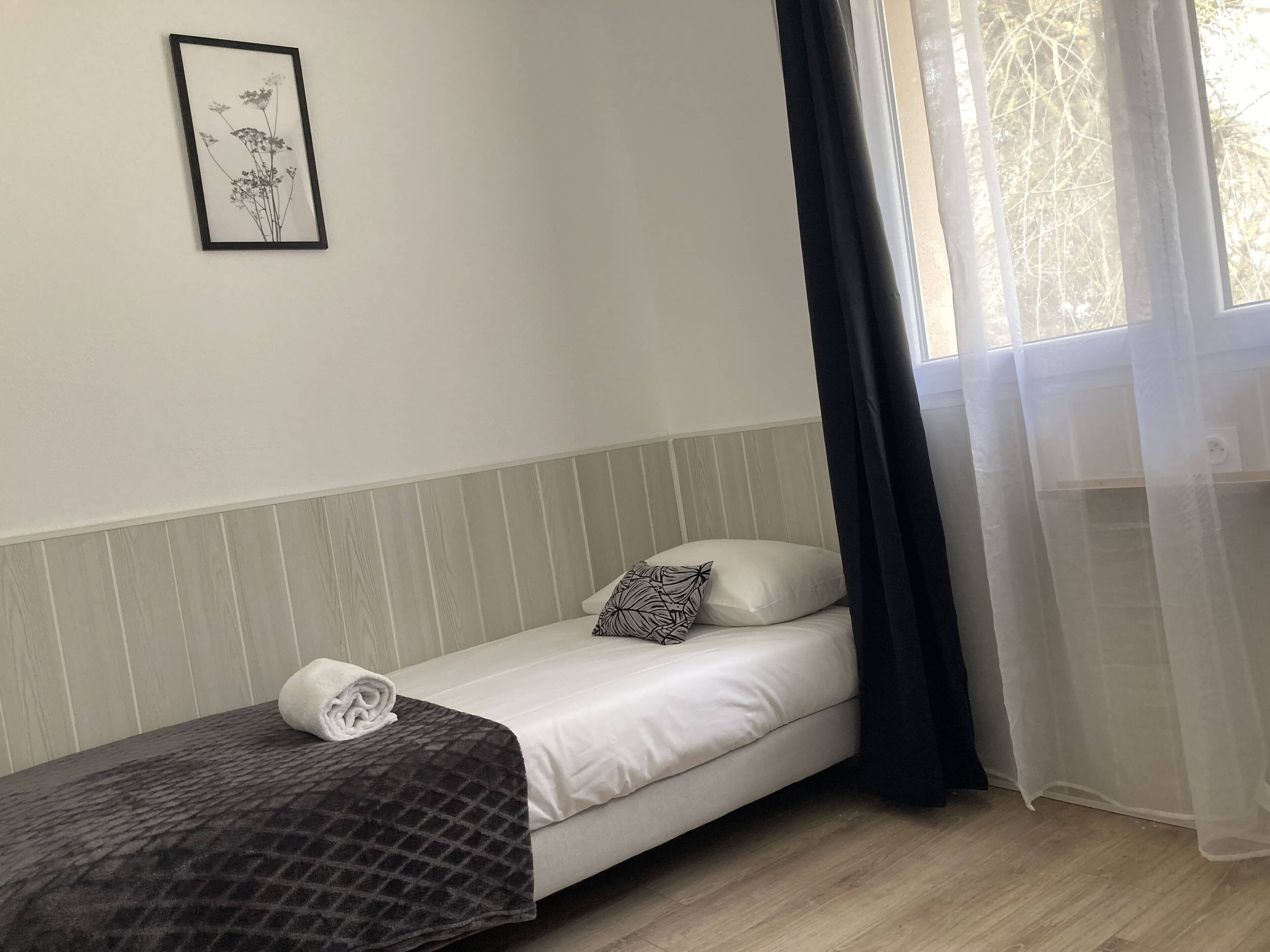 Un letto singolo con un materasso bianco, una coperta scura a motivi e un cuscino con disegno bianco e nero. Sopra il letto è appesa una foto incorniciata.