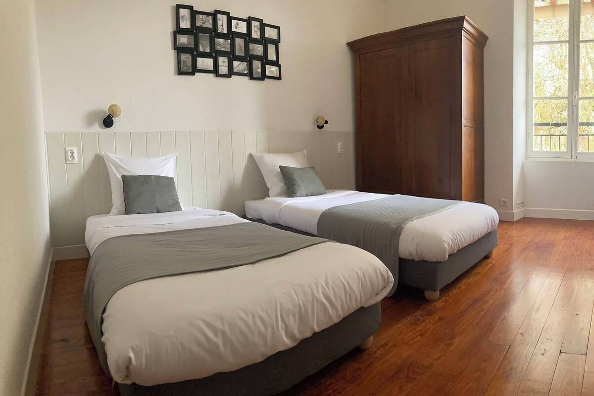 Une chambre avec deux lits, une armoire en bois, un cadre photo fixé au mur et une fenêtre donnant sur des arbres.