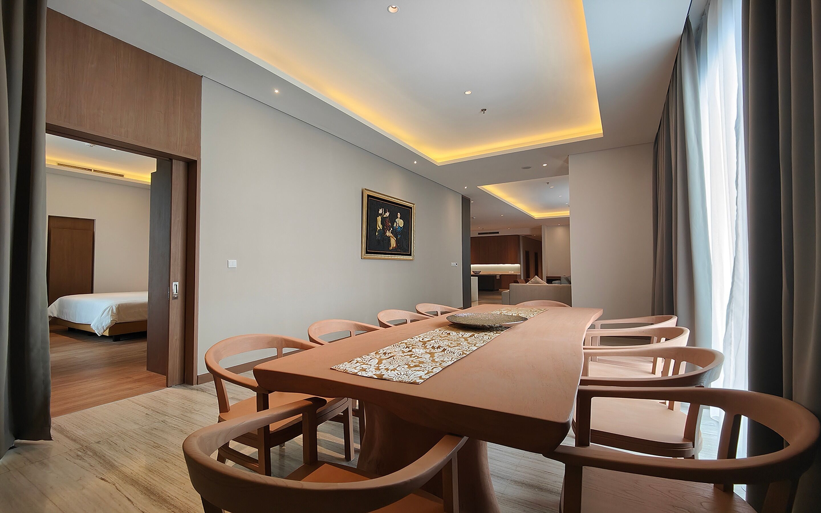 A hotel room with a long dining table, multiple chairs, a bed, and a painting on the wall.