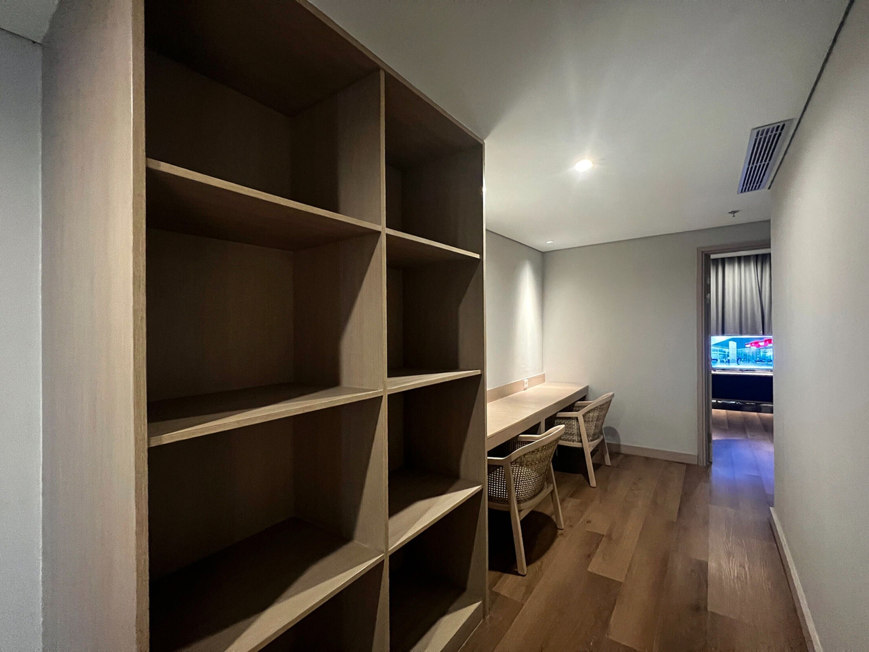 Una habitación moderna con una estantería de madera empotrada, una pequeña mesa con dos sillas y vistas a un paisaje urbano a través de una ventana.