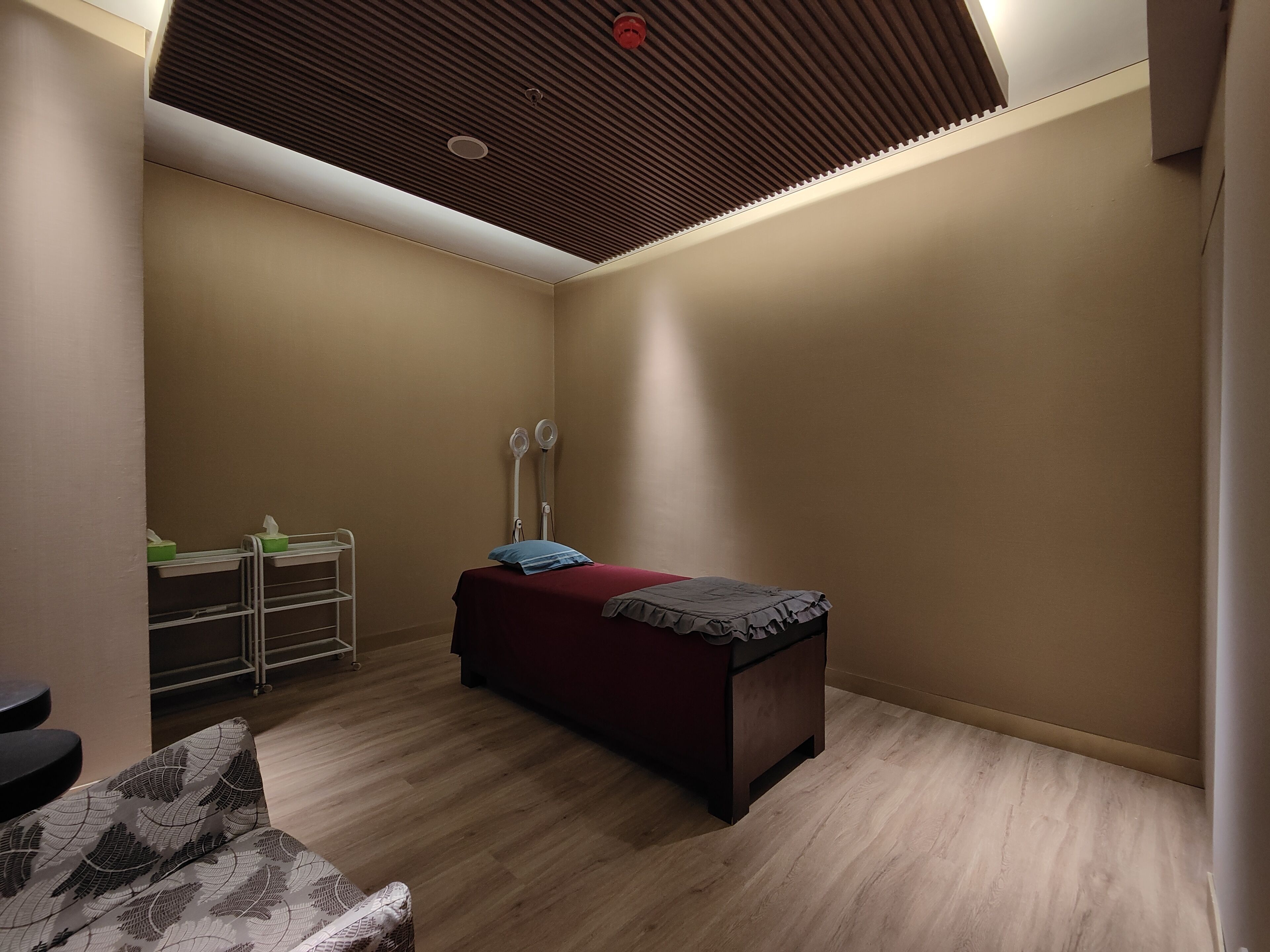 A room with a bed, a small table, a chair, a shelf with items, and a ceiling with a vent.