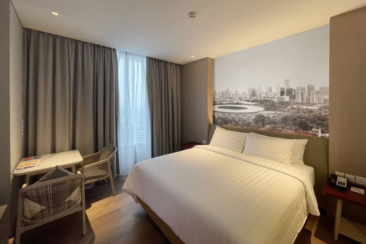 Habitación de hotel con cama, escritorio y silla. Se observa una vista panorámica de la ciudad en la pared.
