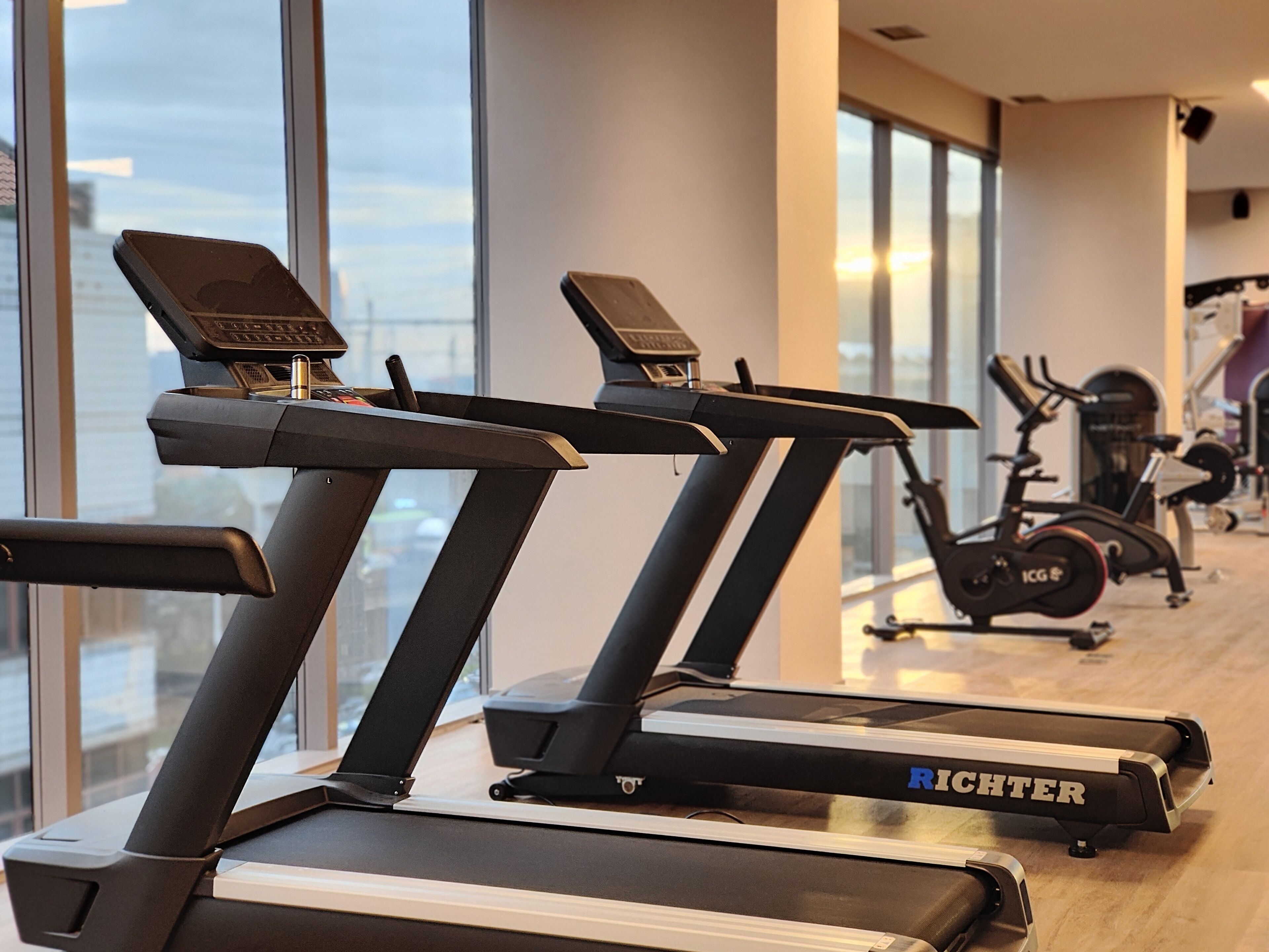 Un gimnasio con cintas de correr y bicicletas estáticas, ventanas amplias y vistas a la ciudad.