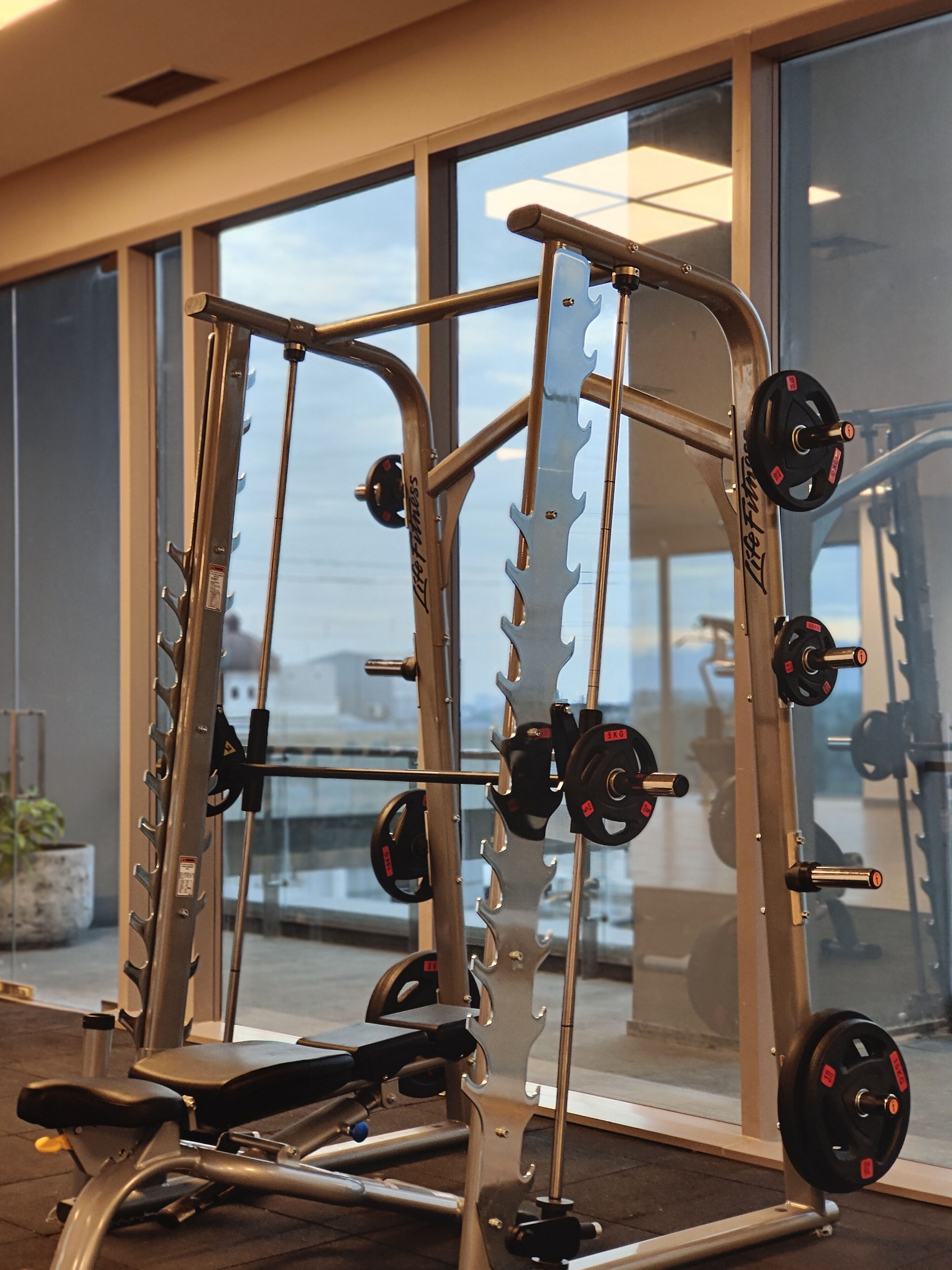A gym with weightlifting equipment, including a bench and a squat rack, with a large window in the background.