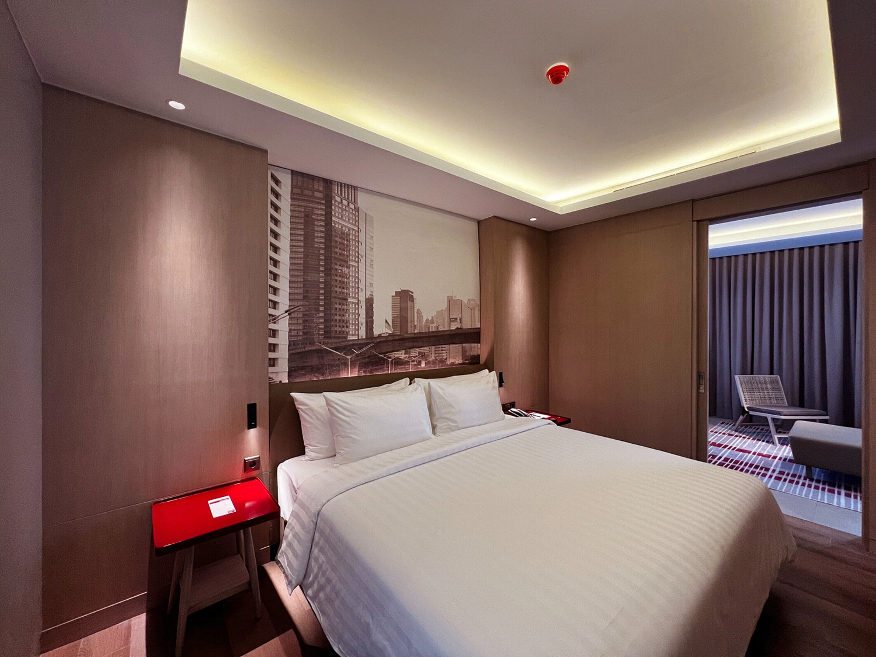 A hotel room with a large bed, a red nightstand, a cityscape mural, and a view of a city through a window.