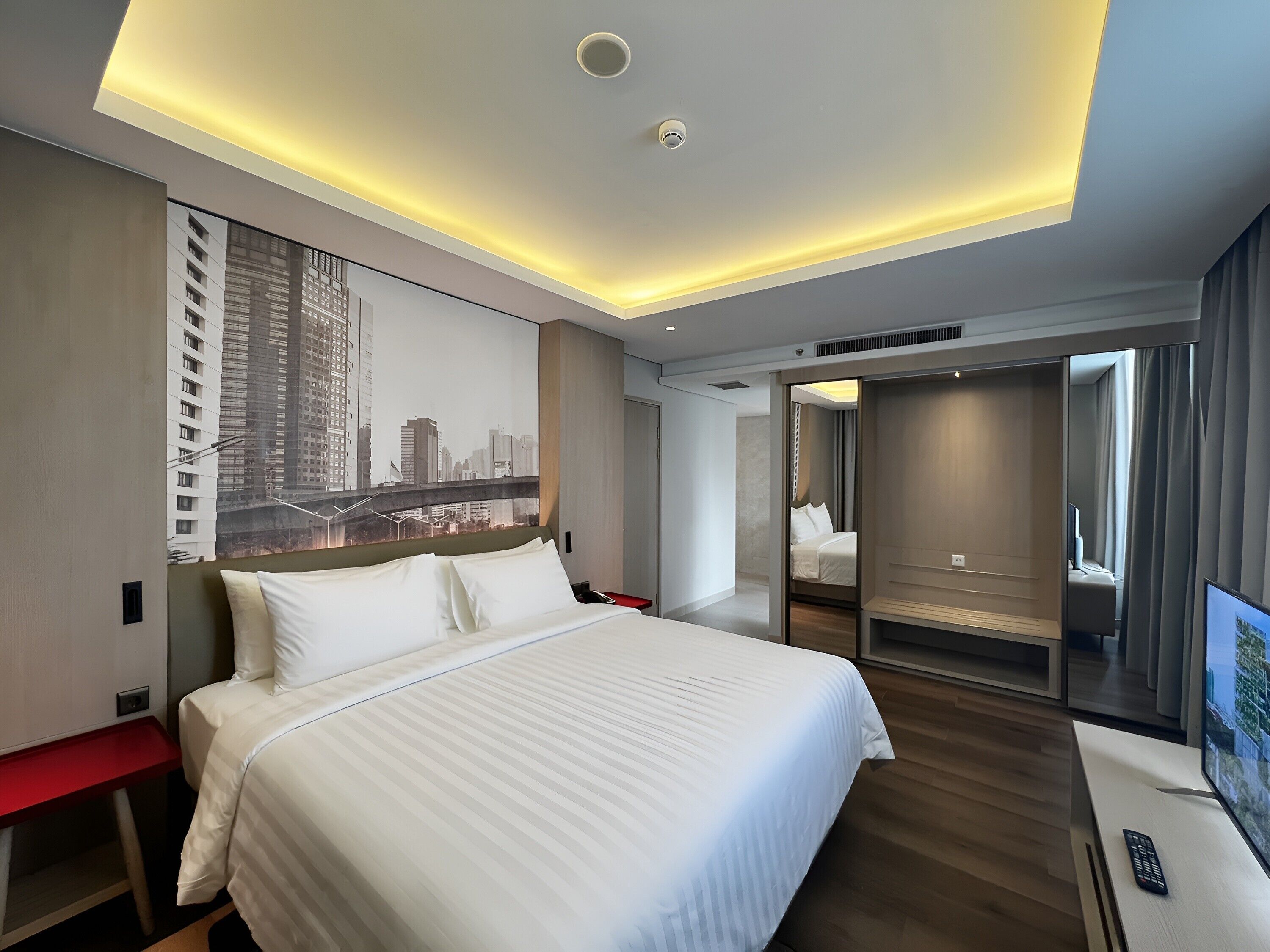 A modern hotel room with a large bed, a cityscape mural, a walk-in closet, and a television.