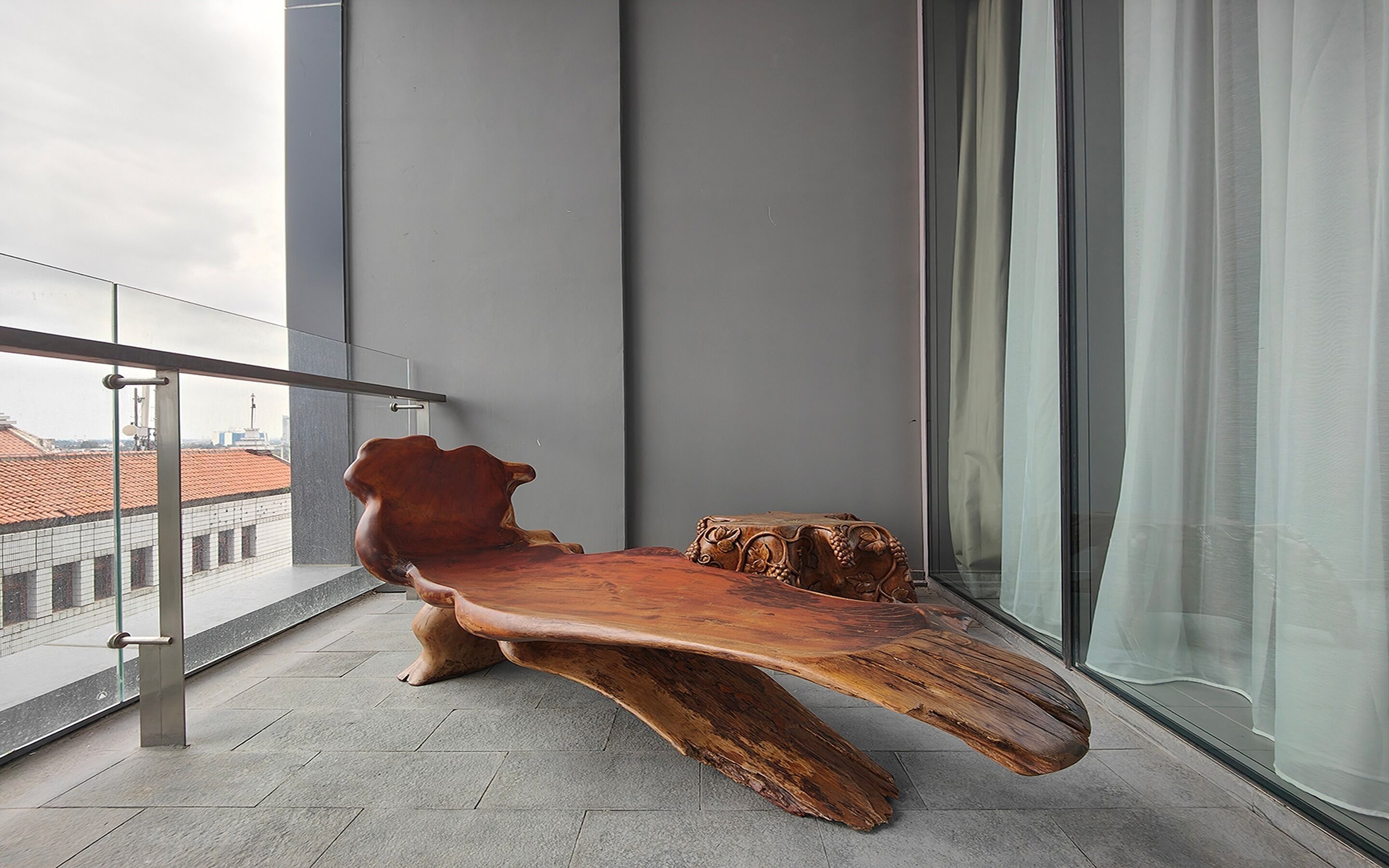 A modern wooden bench with a unique design, positioned on a balcony with a glass railing and a city view.