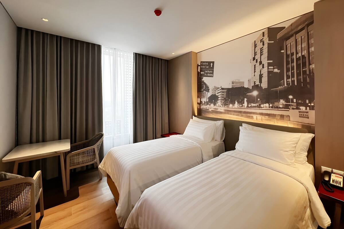 Habitación de hotel con dos camas, un escritorio y una silla. Se observa un cartel con indicaciones y un póster de paisaje urbano en la pared.