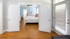 A bedroom with a bed, a dresser, and a nightstand. There is a door leading to another room and a window with blinds. - The Queen B (Oak Island)