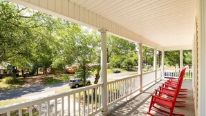 A covered porch with white railings and red chairs, overlooking a street with trees and parked cars. - The Queen B (Oak Island)