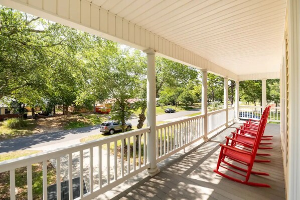 A covered porch with white railings and red chairs, overlooking a street with trees and parked cars.