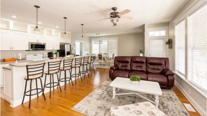 A spacious kitchen with white cabinets, a central island with bar stools, a brown leather sofa, and a ceiling fan. - The Queen B (Oak Island)