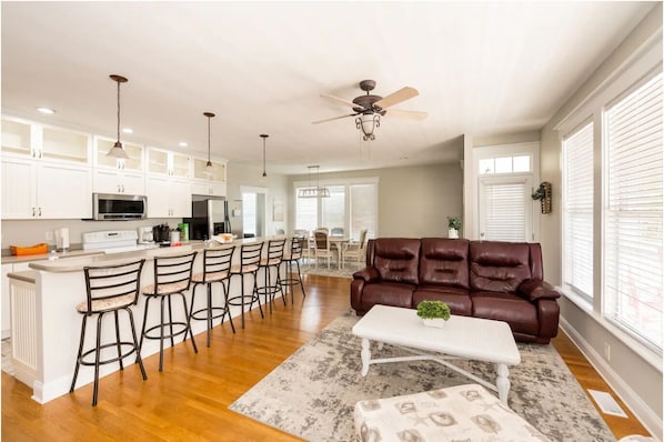 A spacious kitchen with white cabinets, a central island with bar stools, a brown leather sofa, and a ceiling fan.