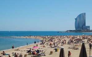Uma praia lotada com pessoas tomando sol e nadando, um veleiro ao longe, e um edifício moderno com design curvo ao fundo.