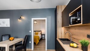 Eine moderne Küche mit integriertem Mikrowellenherd, Spüle und einer Arbeitsplatte mit Zitronen. Ein Essbereich mit Tisch und Stühlen ist vorhanden, und durch einen offenen Durchgang ist ein Schlafzimmer zu sehen.