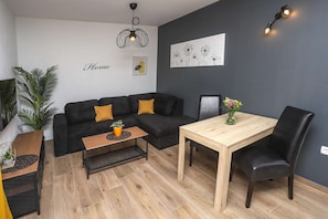 Ein modernes Wohnzimmer mit einem schwarzen Sofa, einem Holztisch, einem Esstisch mit zwei Stühlen, einem Blumentopf und Bildern an der Wand.