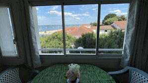 A room with a large window overlooking a beach and buildings. There is a table with a green tablecloth and a vase of flowers. - Beautiful 1-bedroom apartment near the beach, with parking and garage (Arcachon)