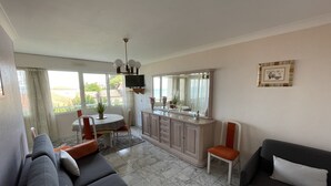 A living room with a sofa, a dining table with chairs, a kitchen area, and a large window with a view. - Beautiful 1-bedroom apartment near the beach, with parking and garage (Arcachon)