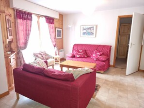 A cozy living room with a red sofa, a wooden coffee table, and a small table with a chair. There is a door leading to another room and a window with curtains.