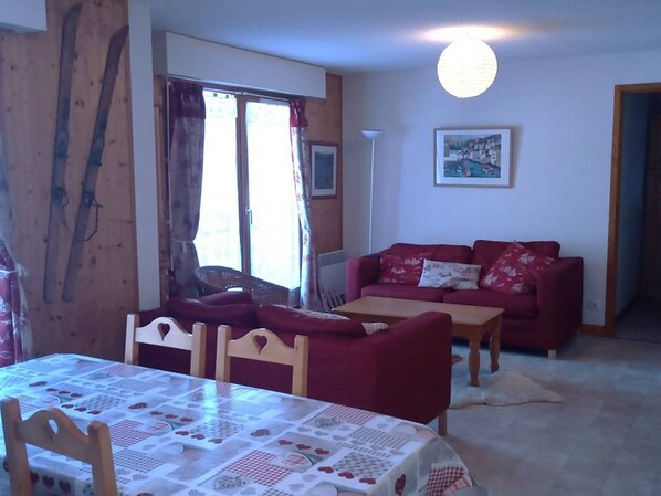 A cozy living room with a red sofa, a wooden dining table, and a large window with curtains. - 3-Room Cabin Rental - Pets Welcome, Parking - At the Foot of Slopes in St Jean d'Aulps (Saint-Jean-d'Aulps)