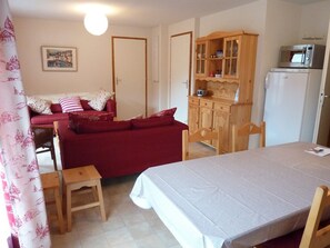 Ett kompakt vardagsrum med en röd soffa, ett matbord i trä och ett köksutrymme med mikrovågsugn och kylskåp.