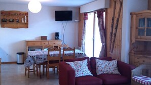 Uma sala de estar acolhedora com um sofá vermelho, uma mesa de jantar de madeira e cadeiras. Uma televisão de tela plana está fixada na parede e há uma janela com cortinas.