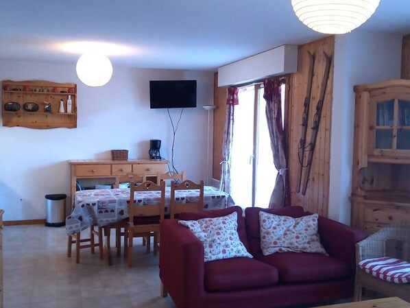 A cozy living room with a red sofa, wooden dining table, and chairs. A flat-screen TV is mounted on the wall, and there's a window with curtains. - 3-Room Cabin Rental - Pets Welcome, Parking - At the Foot of Slopes in St Jean d'Aulps (Saint-Jean-d'Aulps)
