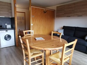 Um espaço de estar compacto com uma mesa de jantar redonda de madeira rodeada por quatro cadeiras, máquina de lavar, sofá de cor escura e armários de madeira.