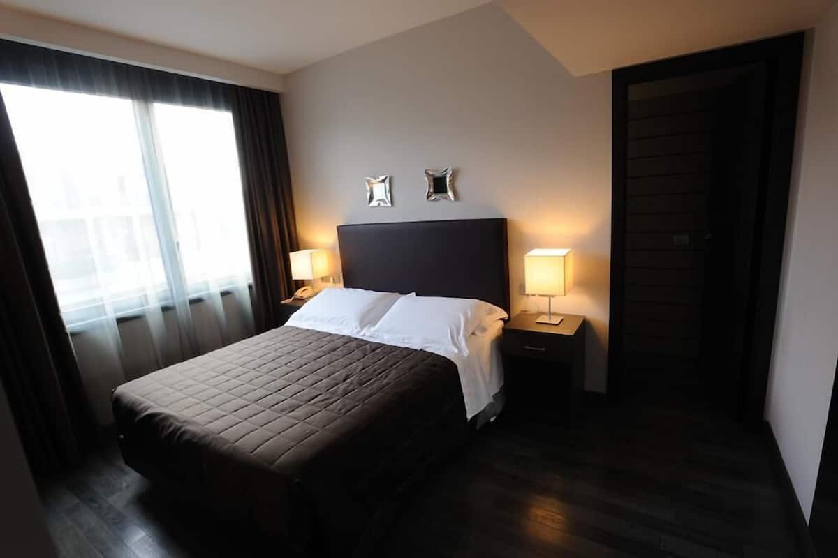 A hotel room with a large bed, two bedside lamps, a dark headboard, and a window with curtains.
