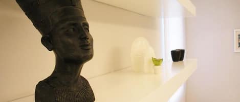 Eine Büstenstatue steht auf einem weißen Regal in einem Raum mit einem weißen Sofa und einem gerahmten Bild an der Wand.