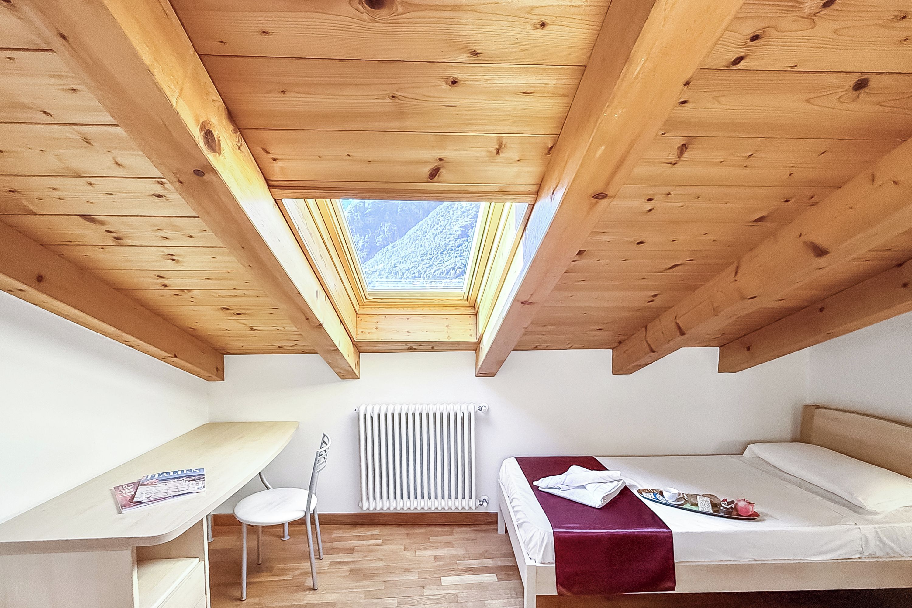 A small, cozy room with a wooden ceiling, a skylight, a bed, a small table, and a radiator.