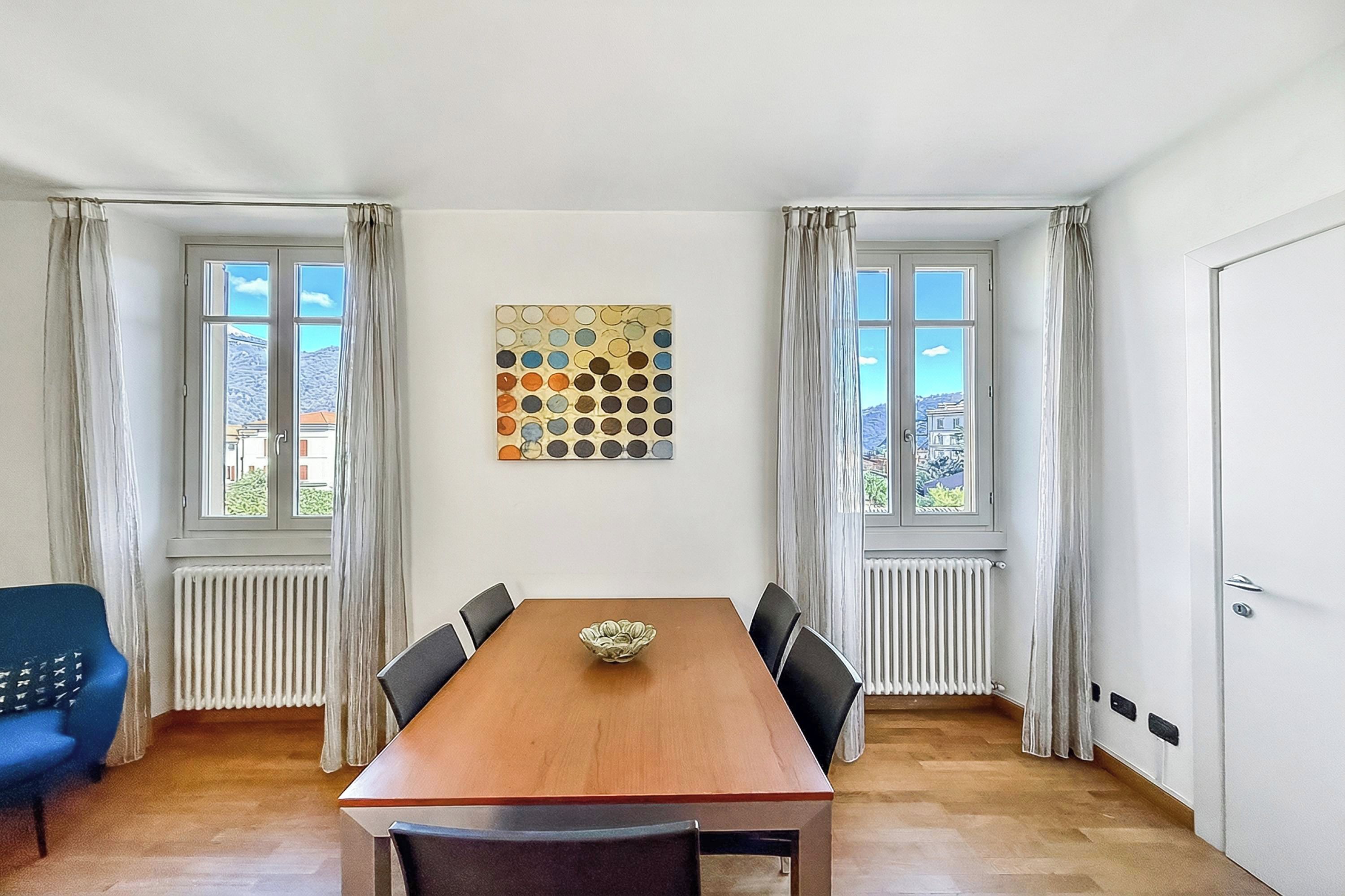 A dining room with a wooden table, six chairs, a decorative wall piece, and two windows with curtains.