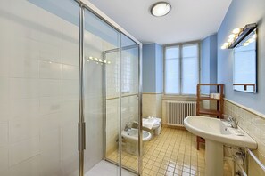 A bathroom with a glass shower enclosure, a toilet, a bidet, a sink, and a mirror with lights.