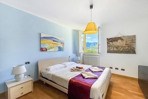 A bedroom with a bed, a bedside table, a lamp, a window with a view, and two wall-mounted artworks.