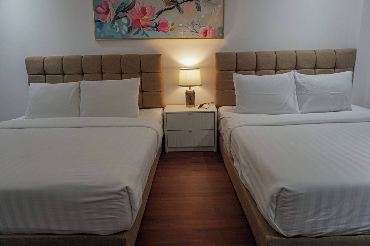 Ein Doppelzimmer mit zwei Betten, einem Nachttisch mit Lampe und einem Blumenbild an der Wand.