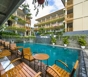 A pool area with wooden lounge chairs and tables, surrounded by balconies with plants and railings. - Graha Socio  Hotel Nusa Dua (Nusa Dua)