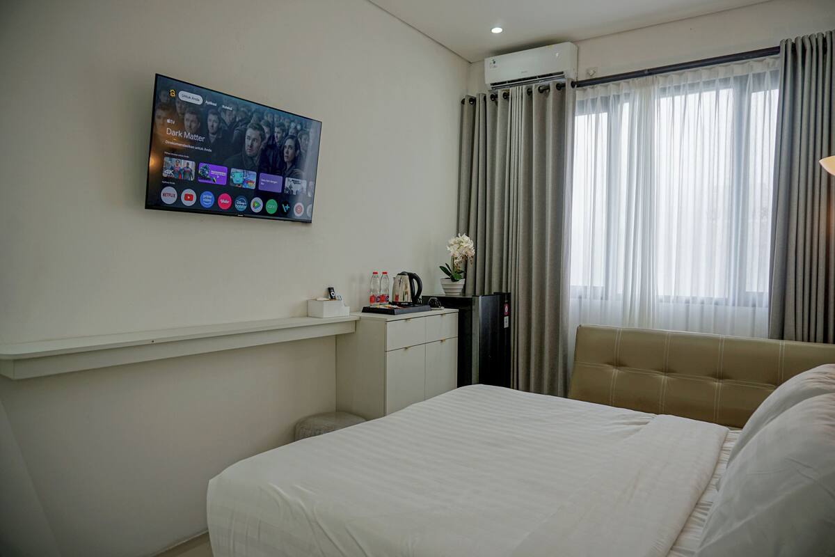Ein Hotelzimmer mit einem Bett, einem Schreibtisch mit Computer, einem an der Wand montierten Fernseher und einem Fenster mit Vorhängen.
