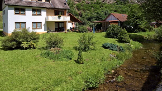 Une maison à deux étages avec un balcon, un petit abri de jardin et un ruisseau au premier plan.
