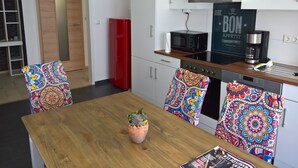 Une cuisine moderne avec des armoires blanches, un réfrigérateur rouge, un four à micro-ondes et une machine à café. On y trouve une table à manger en bois, des chaises colorées et une petite plante, un cactus.