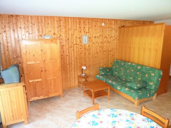 Een kamer met een houten garderobe, een groene bank met bloemenprint, een kleine houten tafel en een ronde tafel met bloemen tafelkleed.