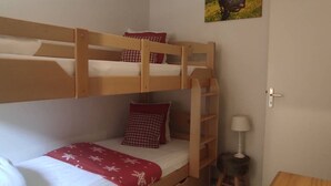 Een slaapkamer met stapelbed, een rood-wit gebedekt matras, een klein krukje, een wandlamp en een afbeelding van een koe op de muur.