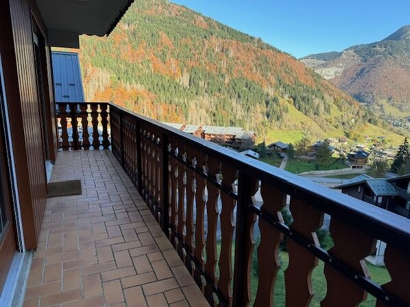 Ein Balkon mit Fliesenboden und Metallgeländer, mit Blick auf eine Berglandschaft mit herbstlich gefärbten Bäumen und ein Dorf darunter.