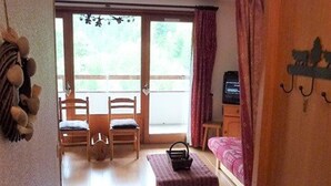 A room with a wooden floor, a bed with a pink blanket, a small table with a basket, two wooden chairs, a TV on a wooden cabinet, and a view through a glass door to an outdoor area. - Studio 28m² with pool, balcony, parking - St-Jean-d'Aulps (Saint-Jean-d'Aulps)