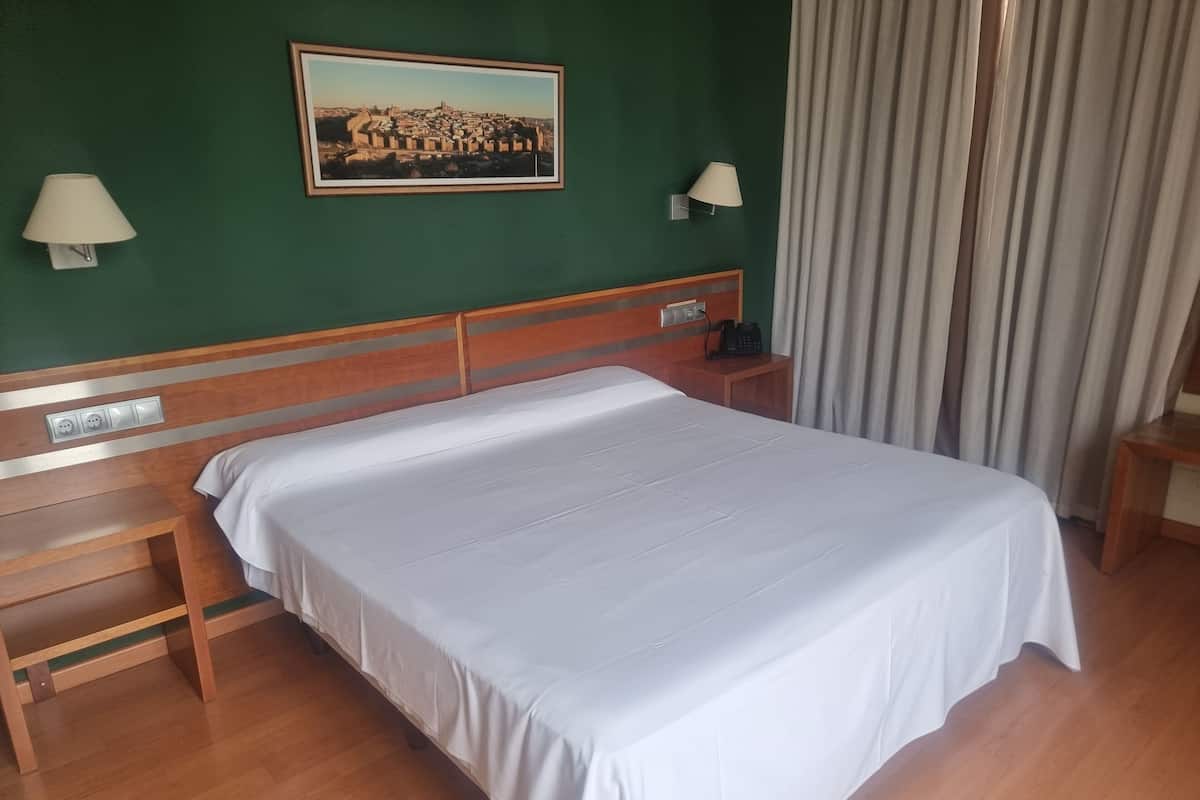 Ein Hotelzimmer mit einem hölzernen Bett, einem Nachttisch, einer Lampe, einem Gemälde an der Wand und einem Vorhang.