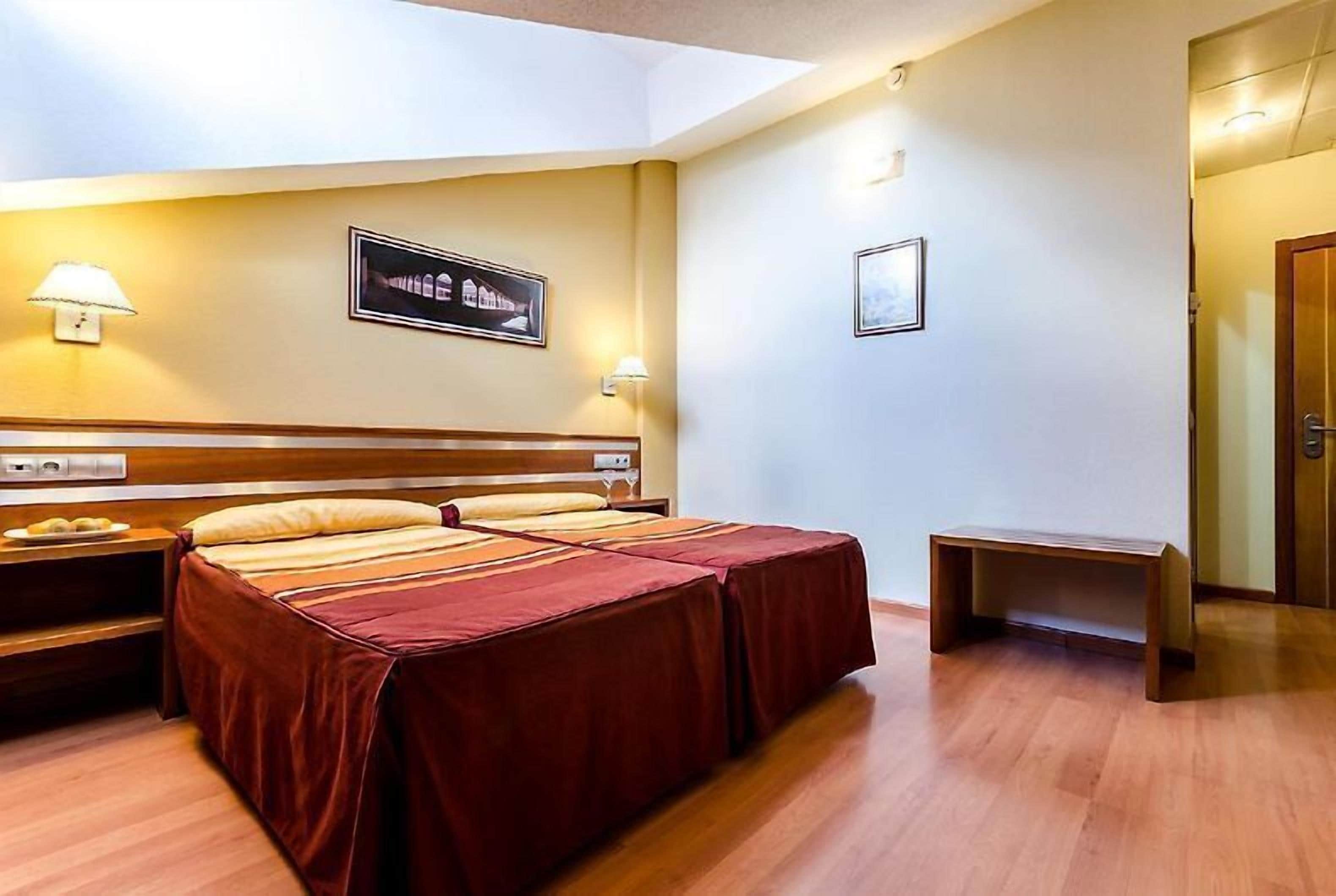 A hotel room with a double bed, bedside tables, a small bench, and wall-mounted lamps.
